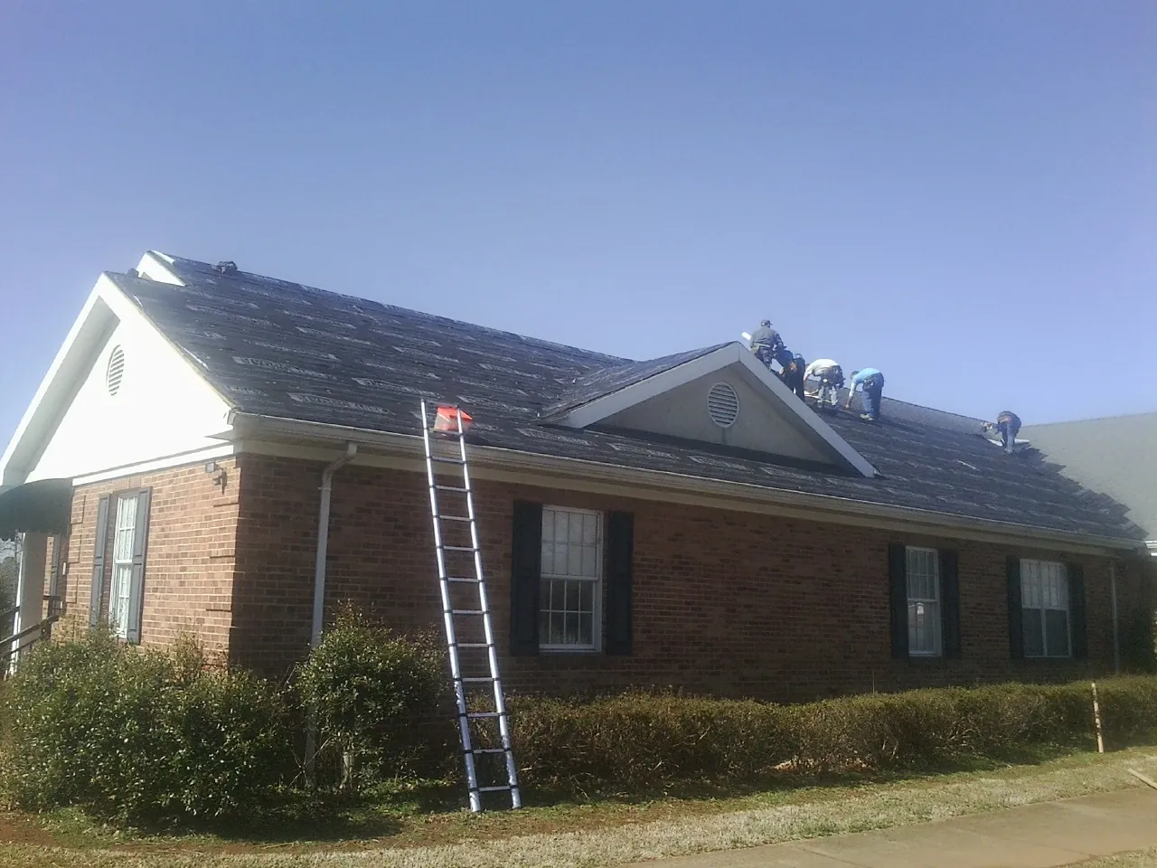 Crew tearing shingles off a brick ranch roof with a ladder set — Emergency Roof Repair work by our crew in Wenham