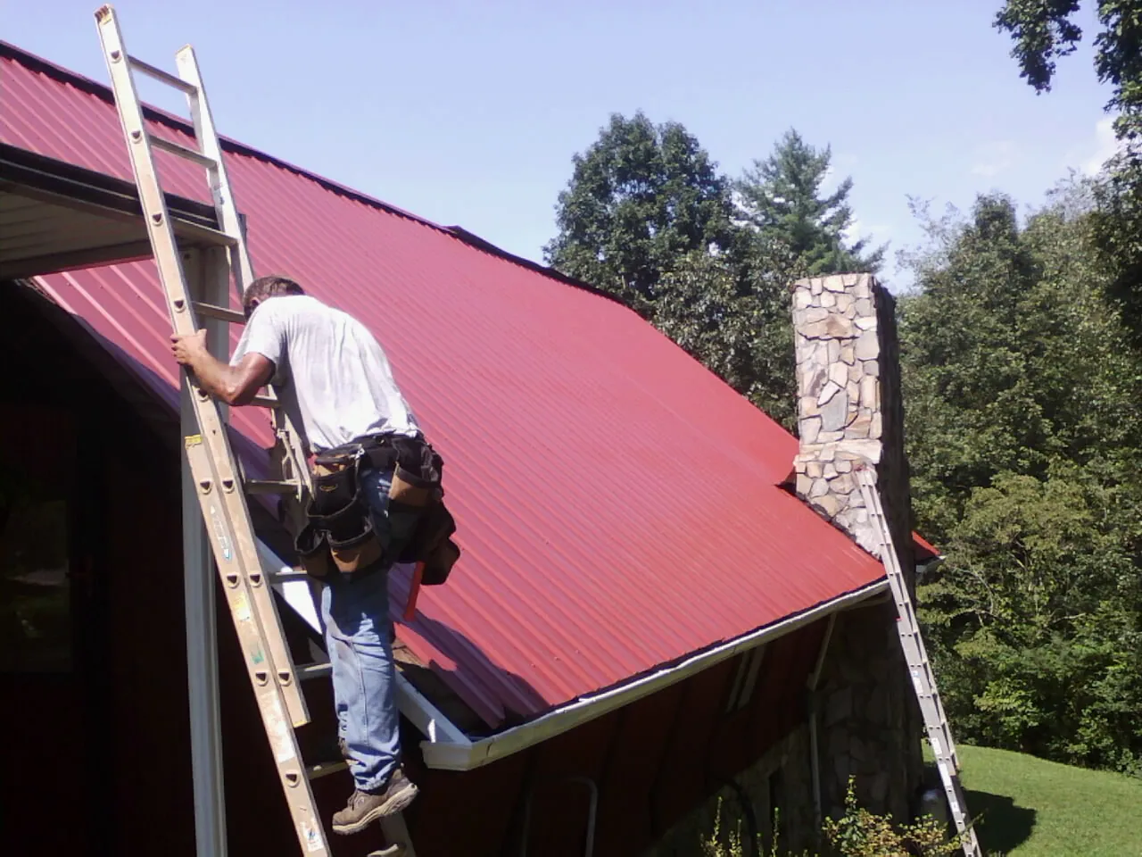 Roofer on a ladder beside a red metal roof and stone chimney — Soffit & Fascia Repair by Roofing Brothers in Wenham