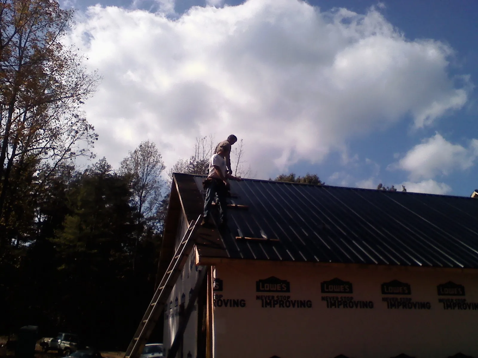 Roofer fastening dark metal panels high against a cloudy sky, from a Roof Inspection job in Wenham
